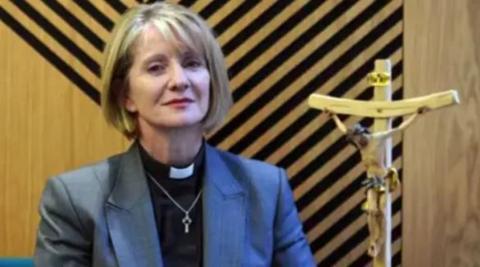 Bishop of Warrington Bev Mason with blond bobbed in front of wooden wall with black stripes on it and wooden figure of Jesus Christ on the cross.
