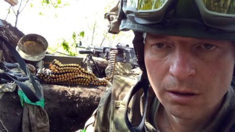 Sergiy Sylantyev, a man in Ukraine's military uniform, with a heavy machine gun and ammunition, taking a selfie.