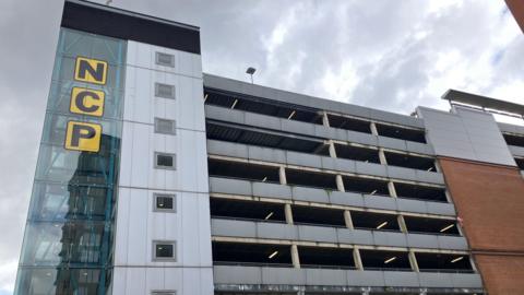 A multi-story car park with brick and glass frontage with the letters N C P on the glass section against the backdrop of a cloudy sky.