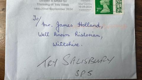 A white envelope with a second-class stamp addressed, handwritten in blue ink, to Mr James Holland, well-known historian, Wiltshire, with the words try Salisbury SP5 added in black ink underneath.