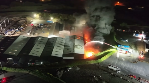 Aerial footage taken at night showing a fire engine spraying water on a fire at a number of sheds at a recycling centre. Smoke can be seen rising from the flames.