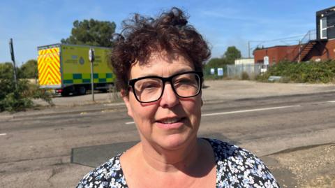 Julie, a woman with short curly hair. she is wearing a floral blue, black and white top, smiling for the camera. Julie is standing on the road, with an ambulance in the background.
