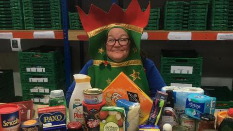 Maria Stevenson, a middle-aged woman in glasses, smiles at the camera while dressed in a Christmas cracker costume that surrounds her face. In front of her on a table is a selection of items: festive biscuits, stuffing mix and other food as well as essentials including toothbrushes and loo roll. There are stacks of green plastic crates behind her.