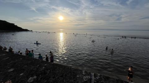 Clevedon Marine Lake. The sun can be seen, low in the sky and reflecting on the water. People can be seen in the area and many in the water.