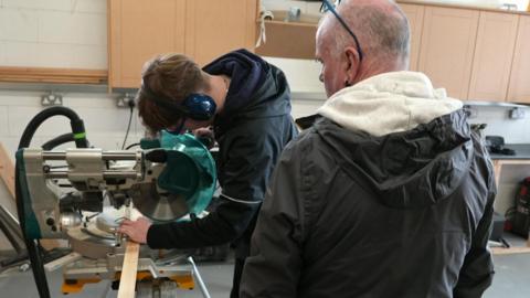 A young person with dark brown hair and wearing a black hoodie operates a circular saw on a workbench. He is overseen by an instructor with a shaved head and wearing a black anorak.