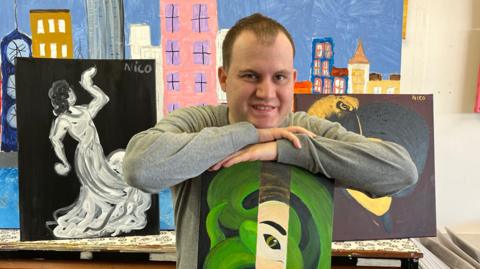 An artist holds a green painting of a snake, in front of other art work he has created. 