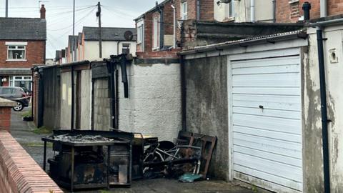 A back alley of a row of houses. There are burnt remains lying on the ground.