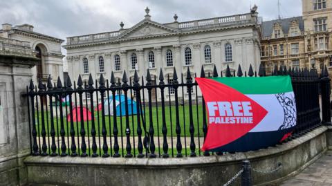 Railings of the Senate House in Cambridge, where tents can be seen on the lawn and a flag has been put on the railings which says FREE PALESTINE