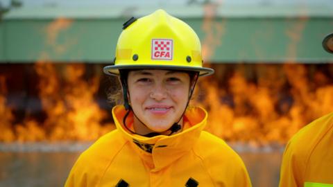 tallarahi n a yellow helmet and yellow firefighting jacket smiling at the camera in front of some fire
