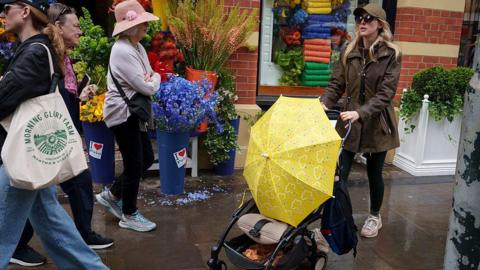 Shoppers passing by a shop displaying flowers on Sloan Square in London. One passer-by is a woman wearing sunglasses and a cap pushing a pram covered with a yellow umbrella. The ground is wet with rain.
