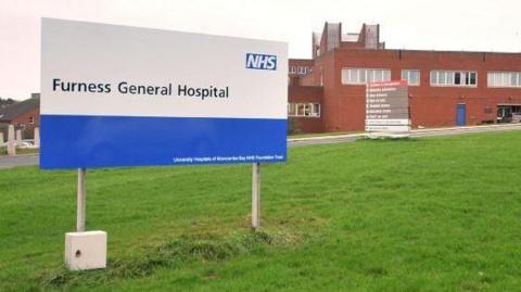 A blue and white sign reading Furness General Hospital is on the grass in the foreground. One of the hospital facilities is at the back. It is a two-storey brown brick building. There is another sign near it with directions to different parts of the hospital. The sky is grey. 