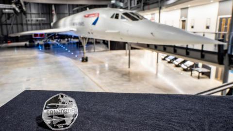 The close up of the coin on a blue surface. The reverse or 'tails' side of the coin portrays the aircraft in flight, against a backdrop of a split-flap departure board, with the word Concorde appearing across the centre. The Concorde plane is in the background.