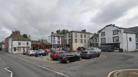 Kirkby Stephen Market Square. Cars are parked in the central paved areas. Shops and pubs surround the square, including one called the Mango Tree.