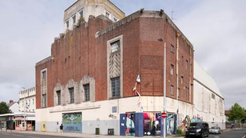 A picture of a large brick and stone building in Plymouth. There are vehicles parked next to it and a main road runs in front of it. There are shops next to the building.