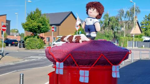 FRIDAY - A red post box with a knitted postbox topper on a residential street in Fareham. The knitting is purple, surrounded by England St George cross flags on a a red string. On top is a knitted football player holding an England flag, with three knitted lions at his feet. Behind the post box is a residential sreet with brick house and metal bollards and street lights on either side of the road.