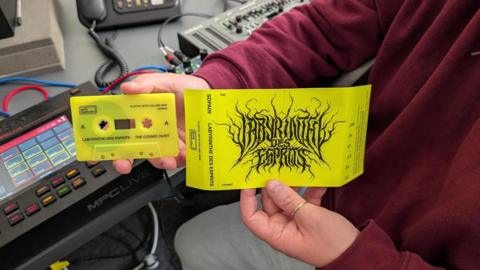 A person wearing a burgundy jumper and grey trousers holds the album cover of an album recorded by mental health unit patients in Devon and a tape of the album. Both the tape and album cover are yellow with black writing on them. The album is called 'Labyrinthe Des Esprits: The Cosmic Hunt'.