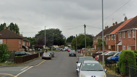 Red brick houses either side of a suburban road, with a number of cars parked on either side of the carriageway, and a cars also parked on driveways