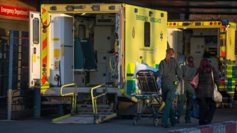 Ambulances with their back doors open parked outside A&E at Gloucestershire Royal Hospital. A group of paramedics are pushing a bed along into the emergency services. 