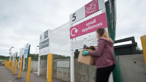 A tip in Oxfordshire. A woman in a red top and blue gloves is getting ready to get rid of her waste.