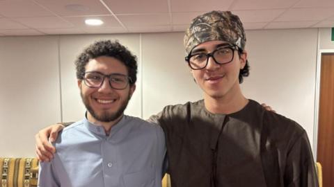 Marwan Ahmed and Mahmoud Elfishawi, two young men standing side by side. Marwan is wearing spectacles, a brown shirt and a patterned bandana around his head, and with his arm around Mahmoud's shoulder. He has dark hair and a close-cropped beard and is wearing a light grey shirt. He is smiling. 