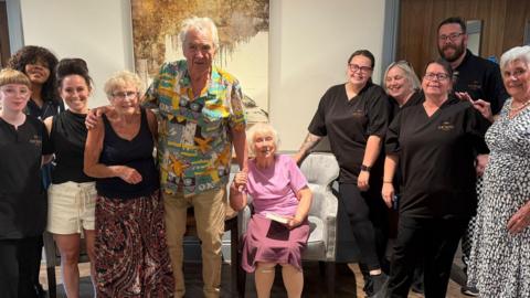 A group of people are stood next to each other with a lady wearing pink sat down in the middle. They are all looking towards the camera smiling. 