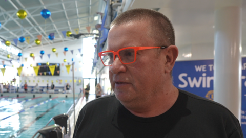 Geof is holding an orange microphone is interviewed beside an indoor swimming pool, with lanes, swimmers, and colourful decorations visible throughout the busy aquatic centre.