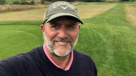 Robert on a golf course, smiling and looking at the camera, wearing a dark jumper with a pink top, with a grey heard and wearing a grey cap. There is grass all around him. 