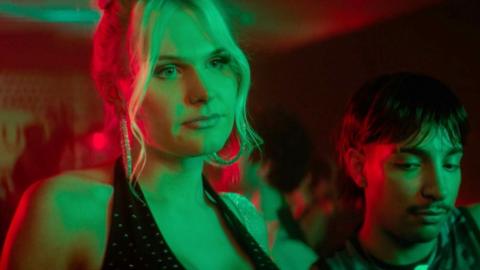 Sasha is wearing a black top. She is in a bar. Green and red lights are shining on her.