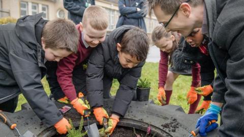 A group of schoolchildren with orange gardening gloves and gardening tools are gathered around a tyre repurposed as a planting area