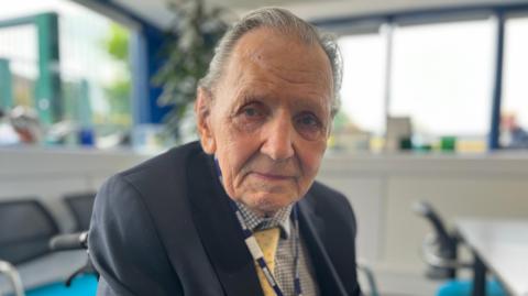 Robert Searles wearing a blue jacket and checked shirt sitting and turning to look at the camera