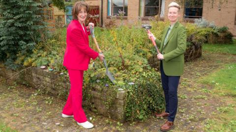 Anna Turner is holding a shovel. She has short brown hair and a pink two piece suit on with white trainers. Stacey Keegan has very short blonde hair, with a green suit jackets and navy trousers. She is also holding a shovel. Behind them is a garden with lots of overgrown plants and weeds 