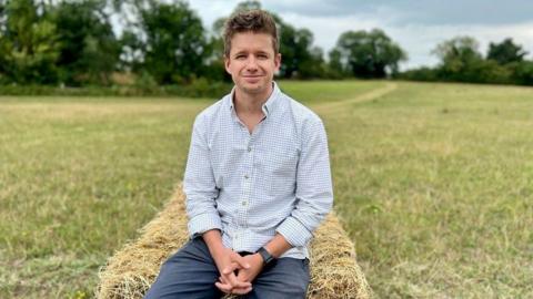 Ed Caudwell smiling for the camera while sitting on a hay bale at his farm. It is a cloudy day.