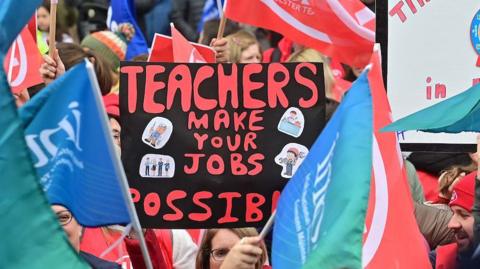 A crowd gathered, one person is holding a sign which says "teachers make your jobs possible"