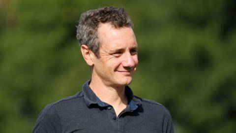 A man with short dark hair smiling and wearing a grey polo shirt.