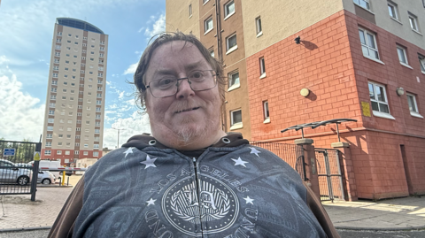 Jamie Lee has ginger hair and a wispy beard, and is wearing a grey hoodie. He is standing in front of tall residential tower blocks. The camera is positioned slightly below him, looking up into his face.