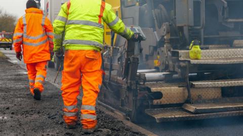 Two men in high-vis jackets and trousers on a construction site. They are facing away from the camera and stood on a gravelly path beside a large van.
