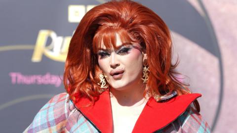 Chanel O'Connor, a drag Queen competing on this year's Ru Paul's Drag Race UK. She is wearing a red wig and lots of makeup. She has in gold earring and is wearing a tartan blazer.