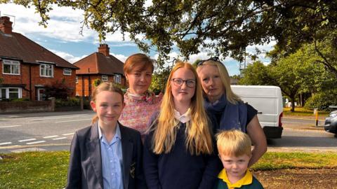 A group of five people look at the camera, they are standing on a patch of grass and next to a road junction. Three of them are children and wearing school uniform, and the two standing at the back are adults.