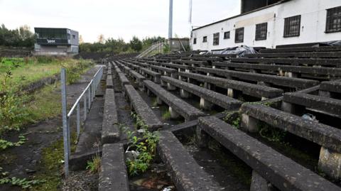 A close-up of the old concrete terraces of Casement, with weeds growing between them. A white building is behind them on the right of the picture and another empty grey two-storey building is on the left.