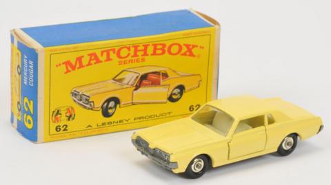 Toy yellow Mercury Cougar car with black wheels. It is on a white backdrop. Behind it is a yellow and blue box that says in red "Matchbox Series" and a picture of the car with the number 62 written in both the left and right hand corners.