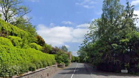 A road through Galashiels with walls and hedges and trees up either side on a sunny day with a few clouds in an otherwise blue sky