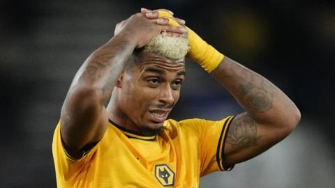 Mario Lemina in gold Wolves shirt and with gold bandage around one wrist places both hands on top of his bleached hair and pulls a pained expression