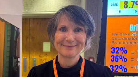 CEO Fiona Brennan stands beside a large “Brighter Futures” charity banner describing support for parents, families and young people in Jersey, inside a brightly lit indoor venue. 