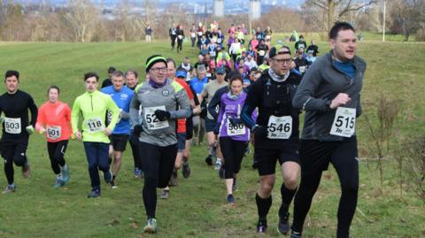 Runners at the Acorn Trails race in Linn Park, Castlemilk, Glasgow on Sunday. There are a number of runners in the image. At the front are three men in dark clothing. A woman in a purple top is behind. They are all mid run. They are in a park with green grass and trees behind them.