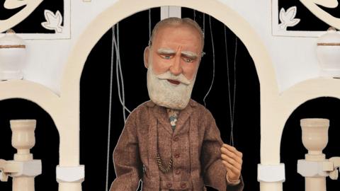 Puppet of a bearded older man in a suit. The puppet, dressed in tweeds, is under an ornate archway.