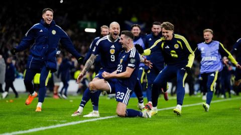 Several Scotland footballers celebrate scoring a goal. The goalscorer has one knee on the turf in celebration, while others rush to congratulate him.