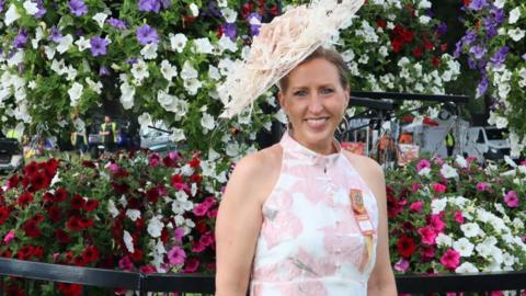 Leanne Garvey wearing a large white floral hat, stood in front of a large display of flowers