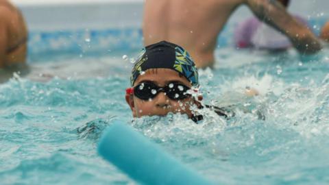 A close-up of a primary age boy swimming in a swimming pool. His chin and mouth are submerged in the blue choppy water and he is wearing black swimming goggles and a black, yellow and blue hat. The torsos of two other people can be seen behind him.
