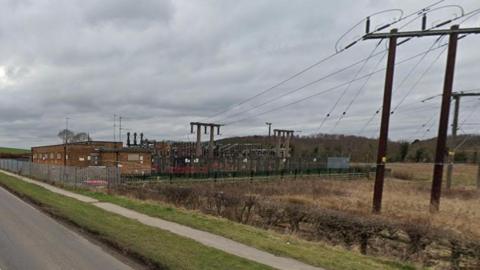 An image of Hard Lane, a road surrounded by fields, with an electricity substation on the right had side of the road.