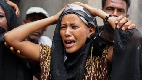 A woman, holding her head, weeps, while a man behind her tries to comfort her